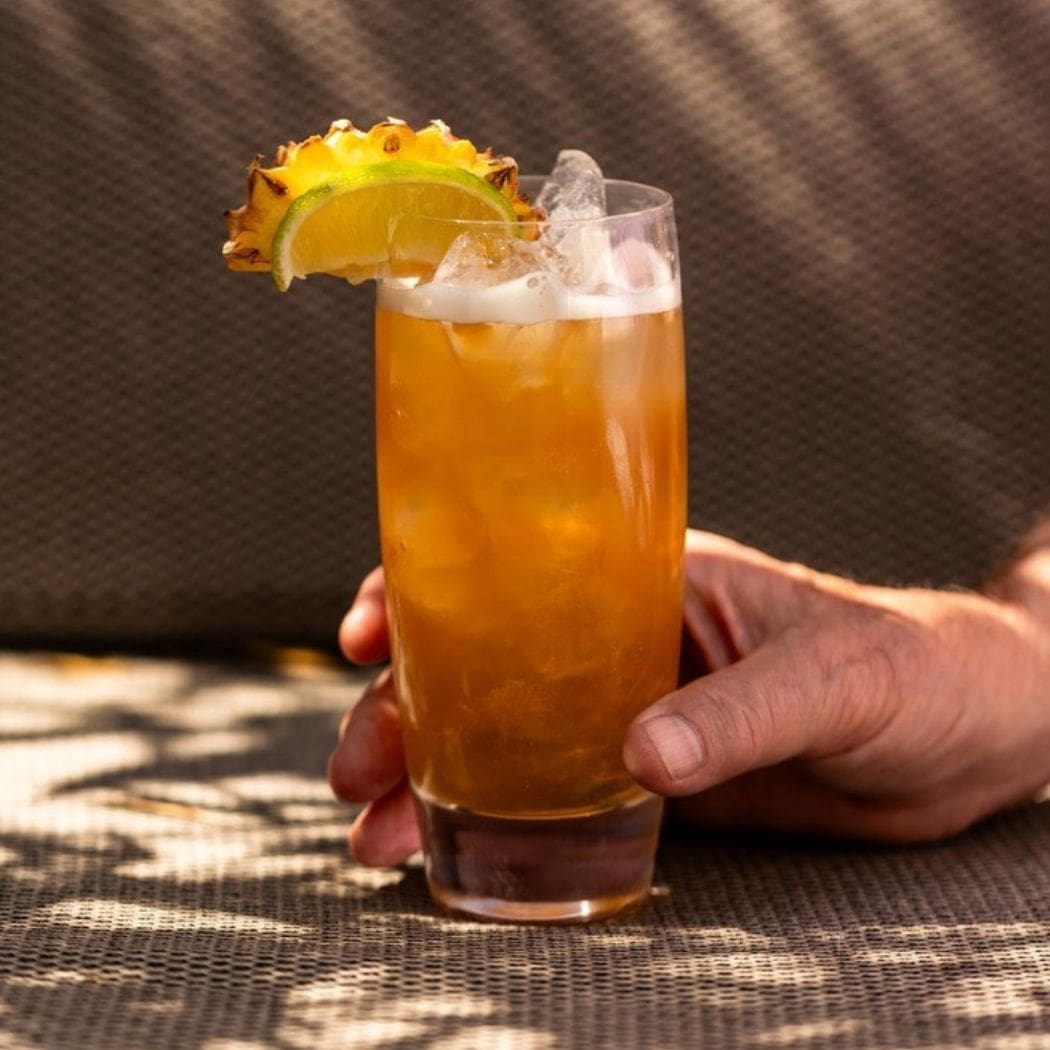 Hand holding a tall iced Rum cocktail with lime and pineapple garnish.