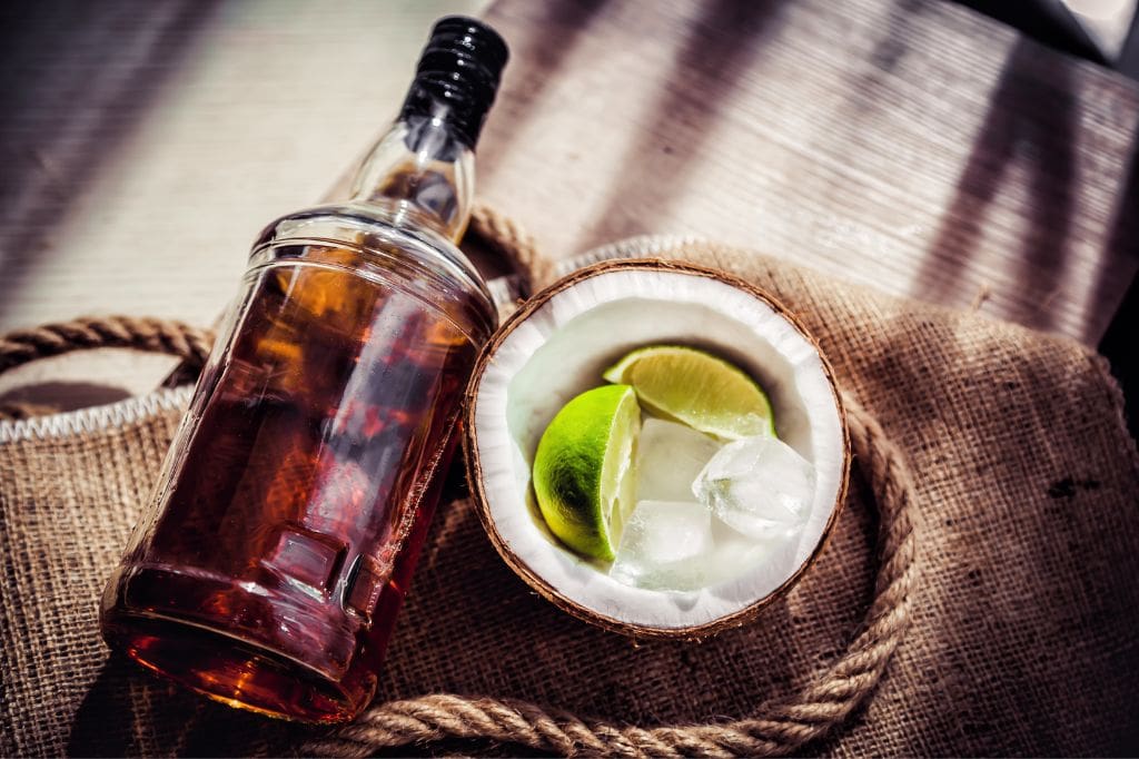 Dark rum bottle beside a coconut half filled with ice and lime wedges.