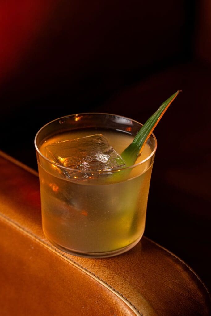 Image 1 alt text: Close-up of a golden clarified pina colada rum cocktail in a clear glass with a large ice cube and a green garnish, set on a brown leather surface in warm ambient lighting.