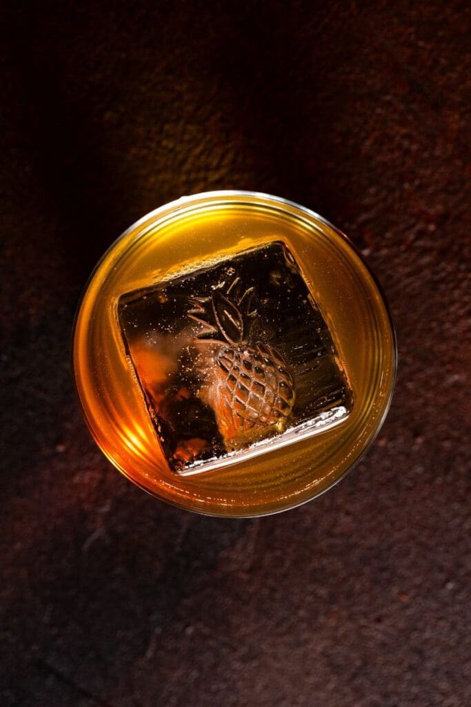 Top view of a golden clarified pina colada cocktail with a large clear ice cube embossed with a pineapple, placed on a dark textured surface under moody lighting.