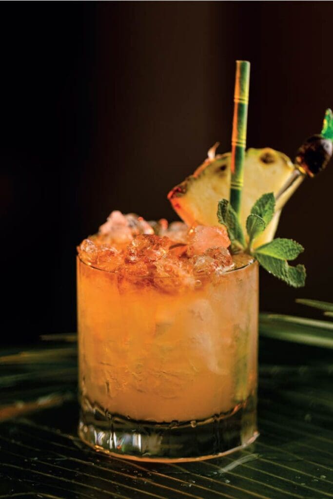 Close-up of a Mai Tai cocktail in a short glass filled with crushed ice, garnished with a pineapple wedge, mint leaves, and a decorative straw, under warm lighting.