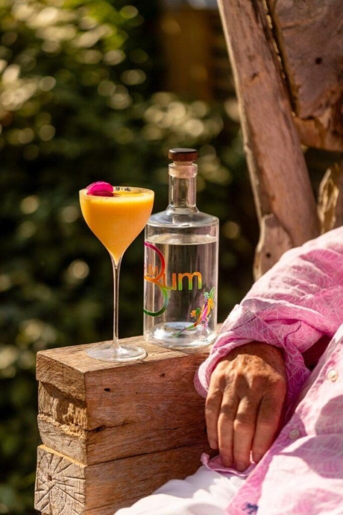 Bottle of craft rum and a tropical cocktail garnished with a flower resting on a rustic wooden bench, with a person in a pink shirt relaxing beside them in an outdoor setting.