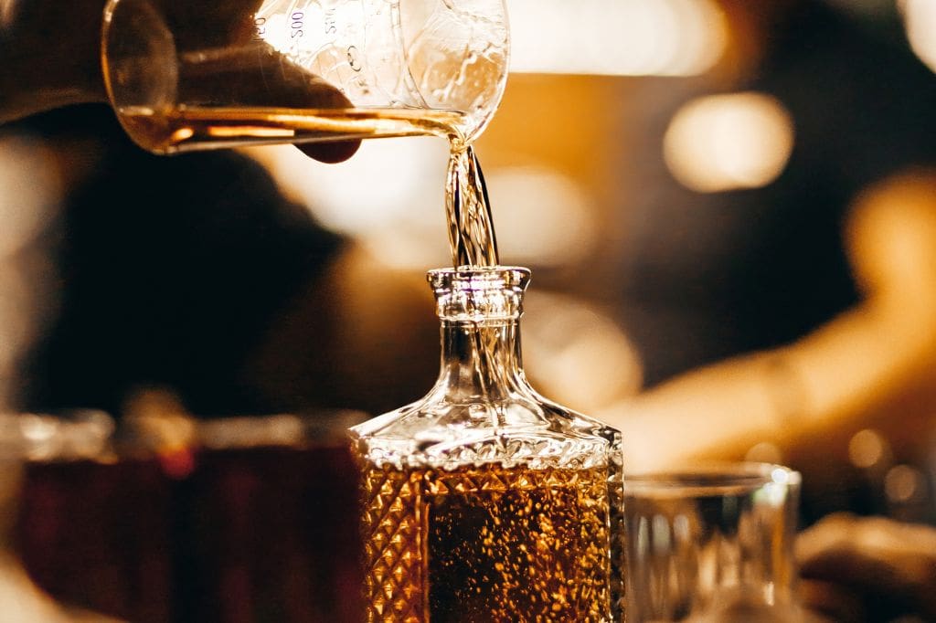 Close-up of amber liquid being poured into a patterned glass decanter, suggesting aged rum in a warm, softly lit bar setting.