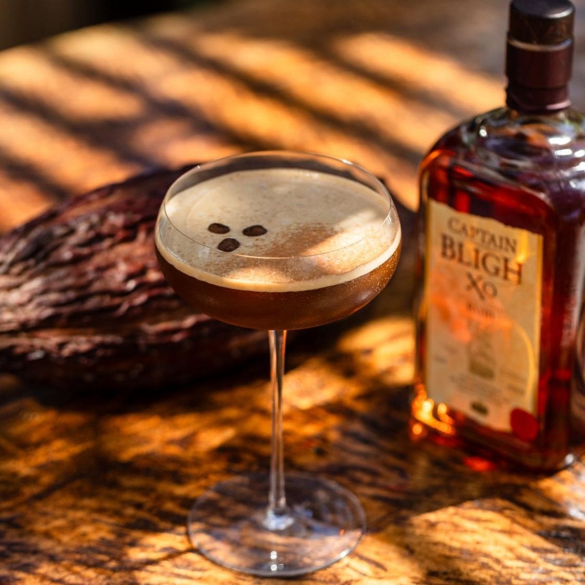 Aged Rum Espresso martini garnished with coffee beans on a wooden surface beside a bottle of Captain Bligh XO aged rum.