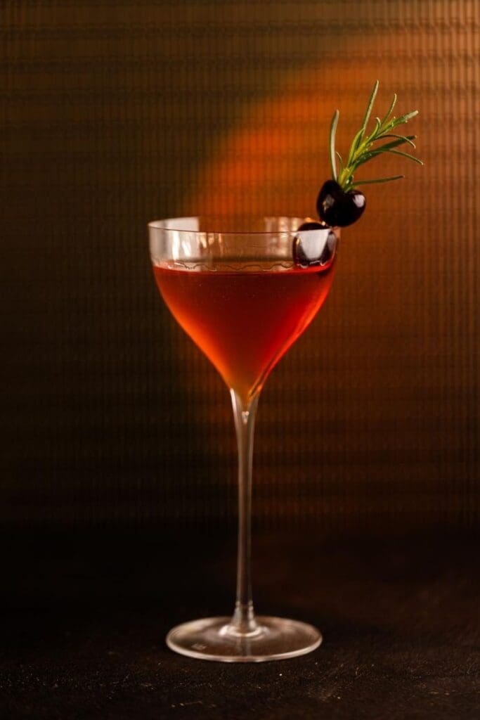 Red rum cocktail in a stemmed glass garnished with cherries and rosemary, set against a warm, moody background.