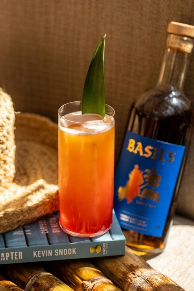 Tall tropical rum cocktail with a pineapple leaf garnish beside a bottle of Basil’s rum, resting on books and surrounded by warm natural textures.