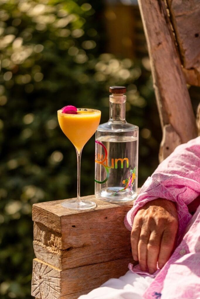 Tropical yellow cocktail with a pink flower garnish beside a bottle of clear craft rum bum white rum, set on a rustic wooden armrest next to a person in a pink shirt.