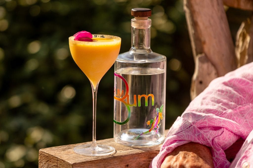 Bright yellow rum cocktail garnished with a pink flower next to a clear bottle labeled "Rum Bum".