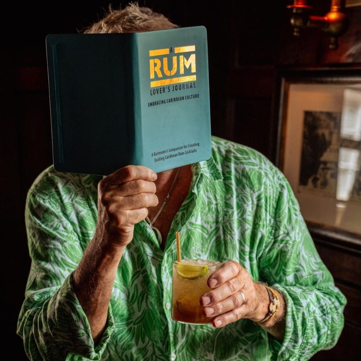 Man in a green tropical shirt holds a cocktail and covers his face with a book titled A Rum Lover’s Journal, ideal for a rum guide.