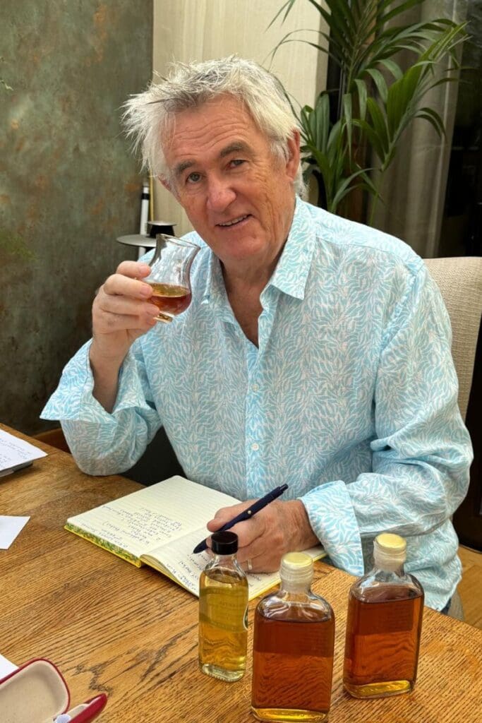 Kevin Snook seated at a wooden table holding a tulip glass of rum liquor while taking tasting notes. Three small bottles of rum sit on the table in the foreground.