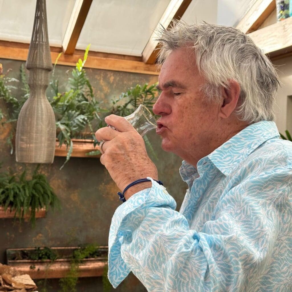kevin Snook the original rum bum in a light blue patterned shirt sips rum from a small tasting glass in a bright indoor garden room with hanging plants.