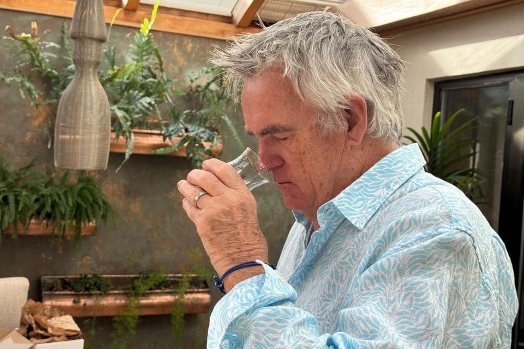 Grey haired man in a light blue patterned shirt smells a small rum tasting glass while standing in a plant filled patio space.