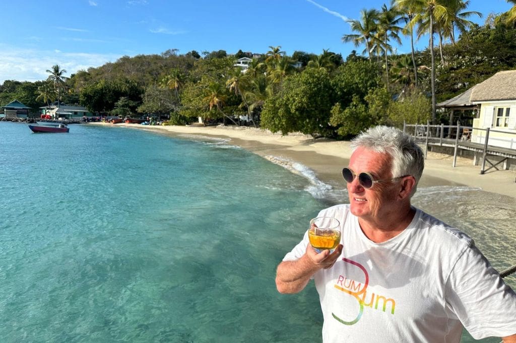 Kevin 'Snooky' Snook, founder of The Original Rum Bum, in sunglasses holding a glass of rum beside clear turquoise water and a palm lined beach.
